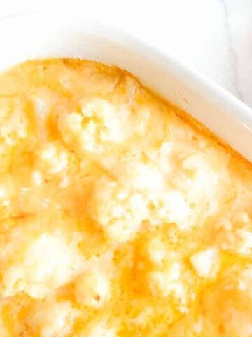 A casserole dish with baked cauliflower in a creamy cheese sauce is placed on a white surface, reminiscent of grandma's cherished recipes. A silver spoon rests on a red and white checkered cloth beside the dish.