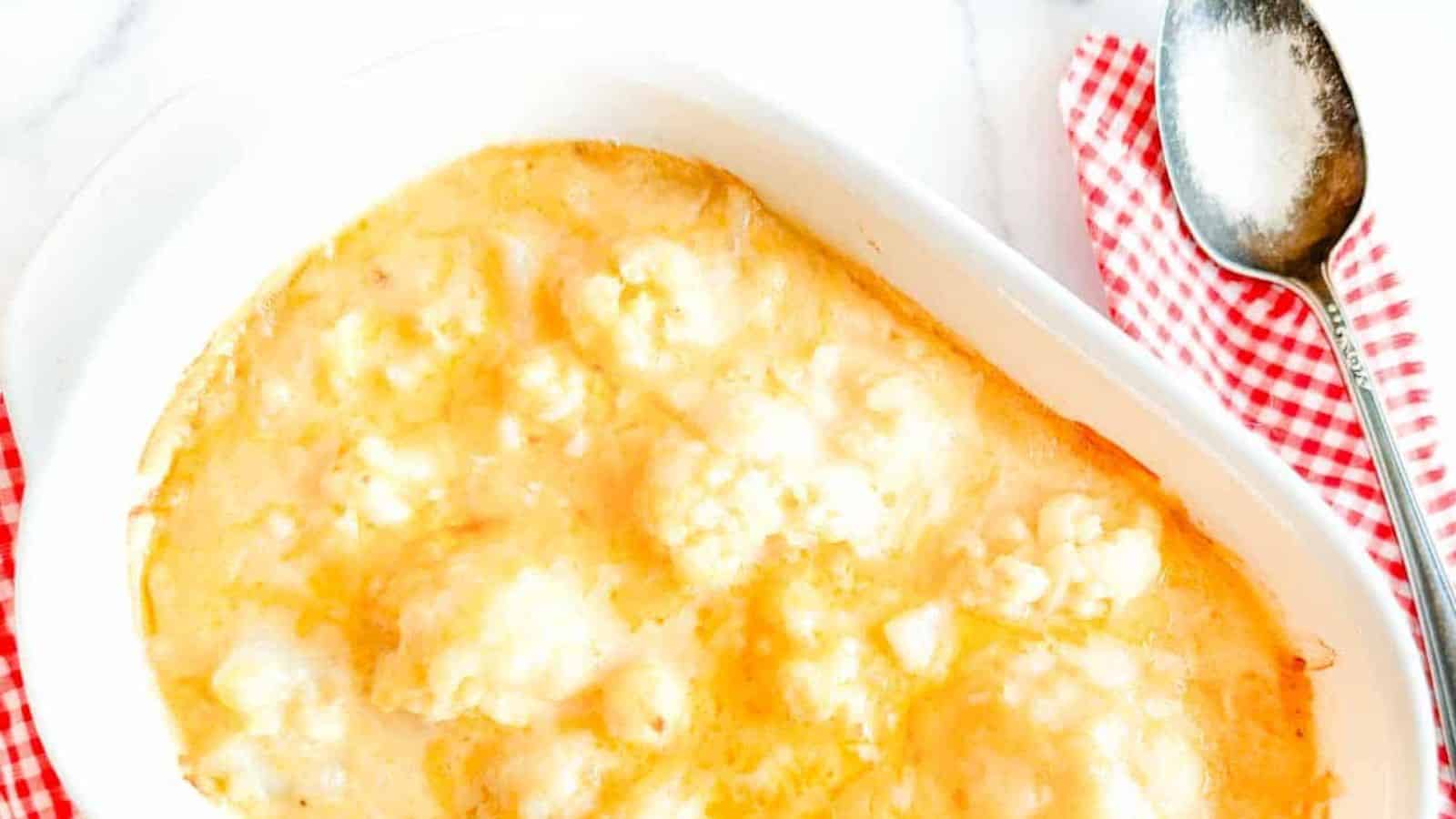 A casserole dish with baked cauliflower in a creamy cheese sauce is placed on a white surface, reminiscent of grandma's cherished recipes. A silver spoon rests on a red and white checkered cloth beside the dish.