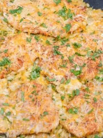 A skillet containing baked chicken breasts topped with melted cheese and chopped parsley, resting on a bed of seasoned rice mixed with vegetables.