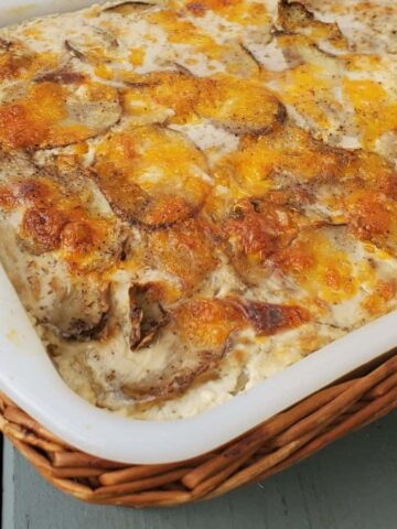 This comfort dish, an instant favorite, is a baked casserole in a rectangular white dish with a wicker holder on a wooden surface. The top is perfectly browned and sprinkled with sliced potatoes and cheese, making it an inviting addition to your collection of go-to recipes.
