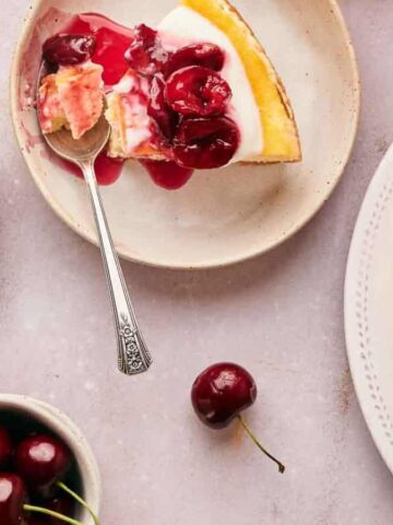 Plates of cherry-topped cheesecake are arranged on a light surface. A slice with a spoon rests on one plate, while a whole cheesecake and a small bowl of cherries are nearby. A single cherry lies on the table.