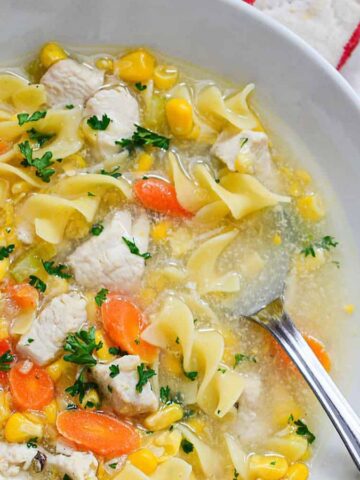 A bowl of warm chicken noodle soup, featuring tender pieces of chicken, egg noodles, carrots, corn, and parsley with a spoon in it. This comforting meal sits on a marble countertop next to a fresh bread roll.