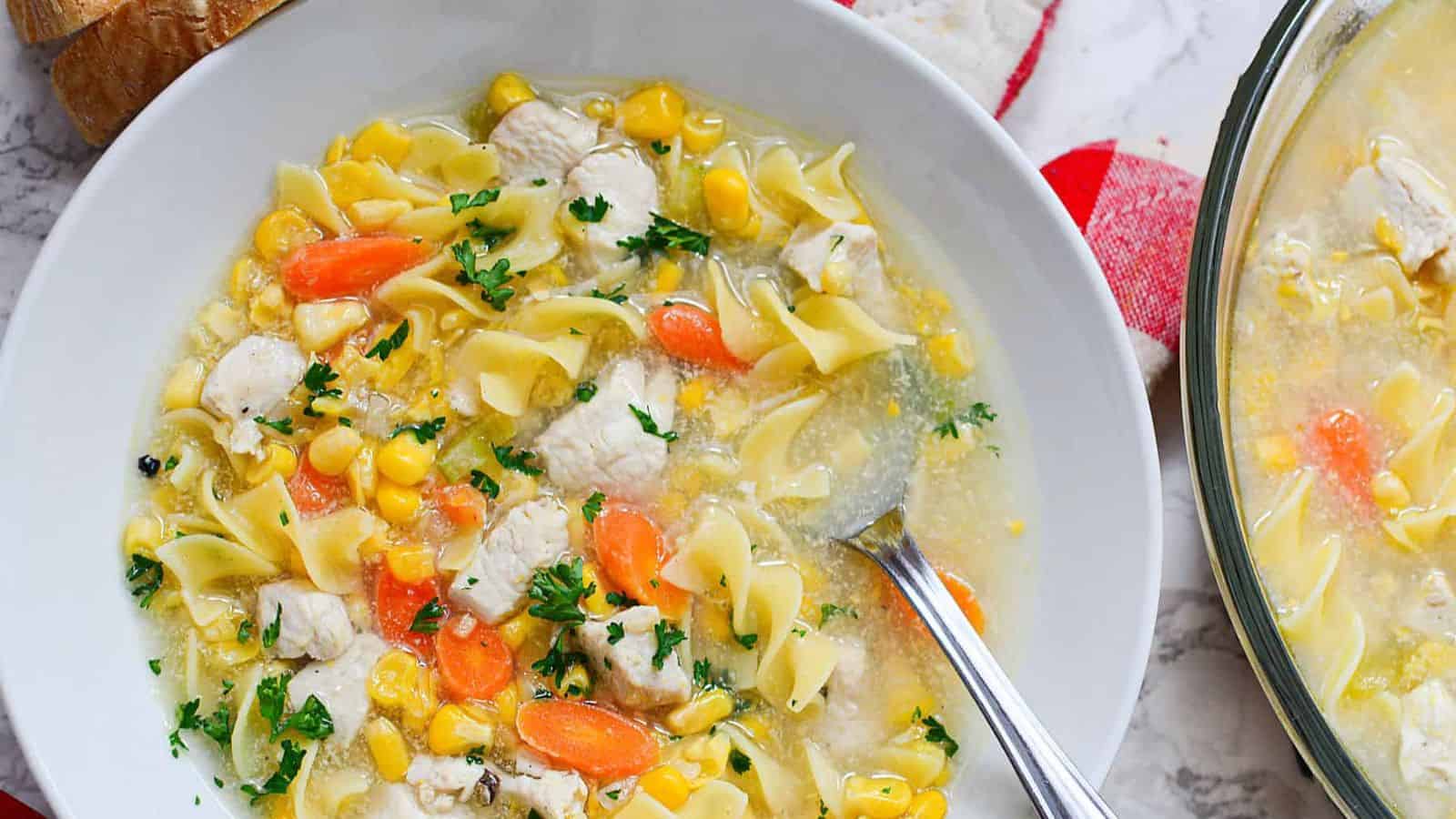 A bowl of warm chicken noodle soup, featuring tender pieces of chicken, egg noodles, carrots, corn, and parsley with a spoon in it. This comforting meal sits on a marble countertop next to a fresh bread roll.