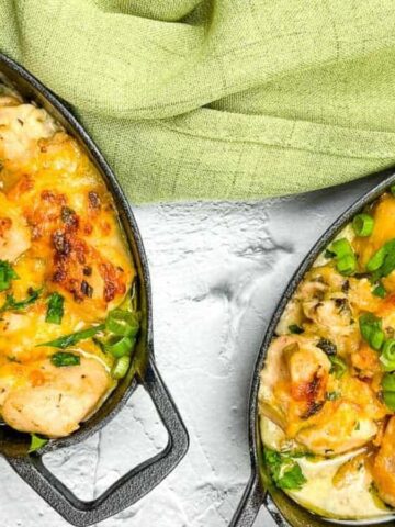 Two black oval dishes filled with a baked dish of chicken, cheese, and green vegetables, garnished with chopped herbs. A light green cloth is draped above the dishes. The white textured surface adds contrast to the presentation.