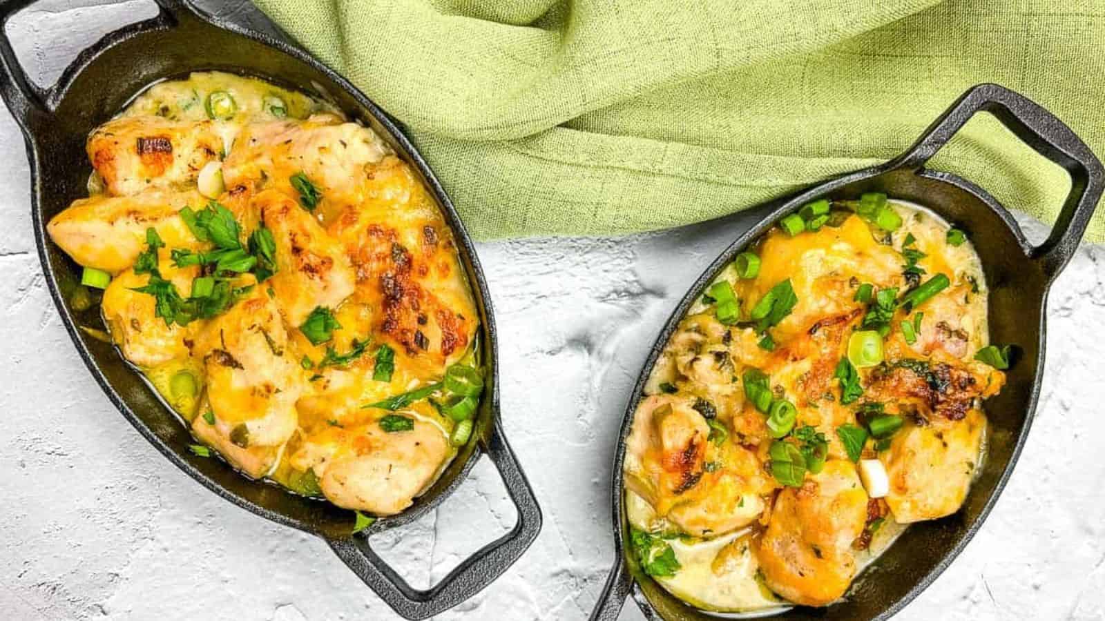 Two black oval dishes filled with a baked dish of chicken, cheese, and green vegetables, garnished with chopped herbs. A light green cloth is draped above the dishes. The white textured surface adds contrast to the presentation.