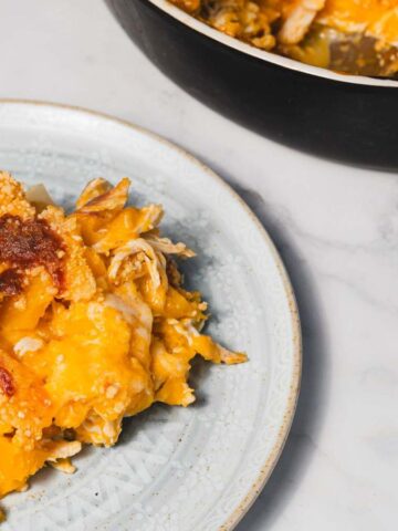 A plate of cheesy chicken casserole is served on a light-colored plate with a fork beside it. The casserole has a golden-brown crust and appears creamy. Part of the casserole dish is visible in the background on a marble countertop.