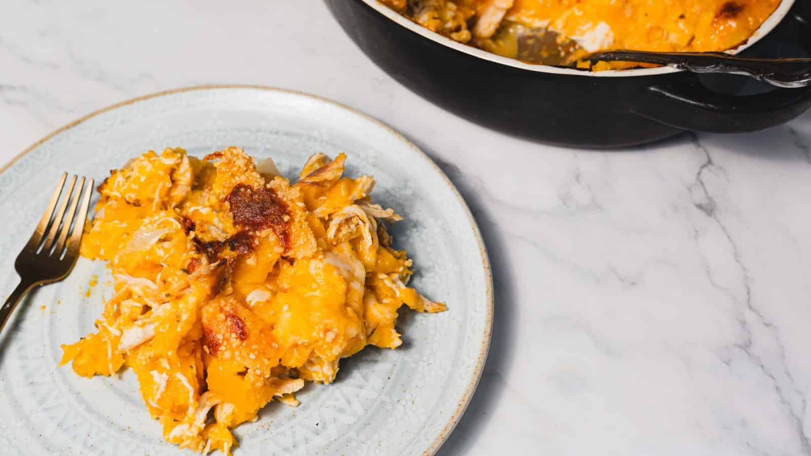 A person lifts a spoonful of cooked macaroni and cheese from an oval black baking dish. The dish is topped with a breadcrumb crust, and a blue cloth is partially visible underneath. The table surface is white.