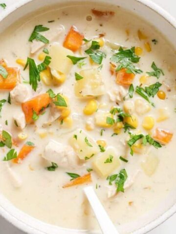 A bowl of creamy chicken soup containing diced carrots, corn, potatoes, and herbs. The soup is garnished with chopped parsley and has a white spoon in it. The bowl is placed on a light-colored surface.