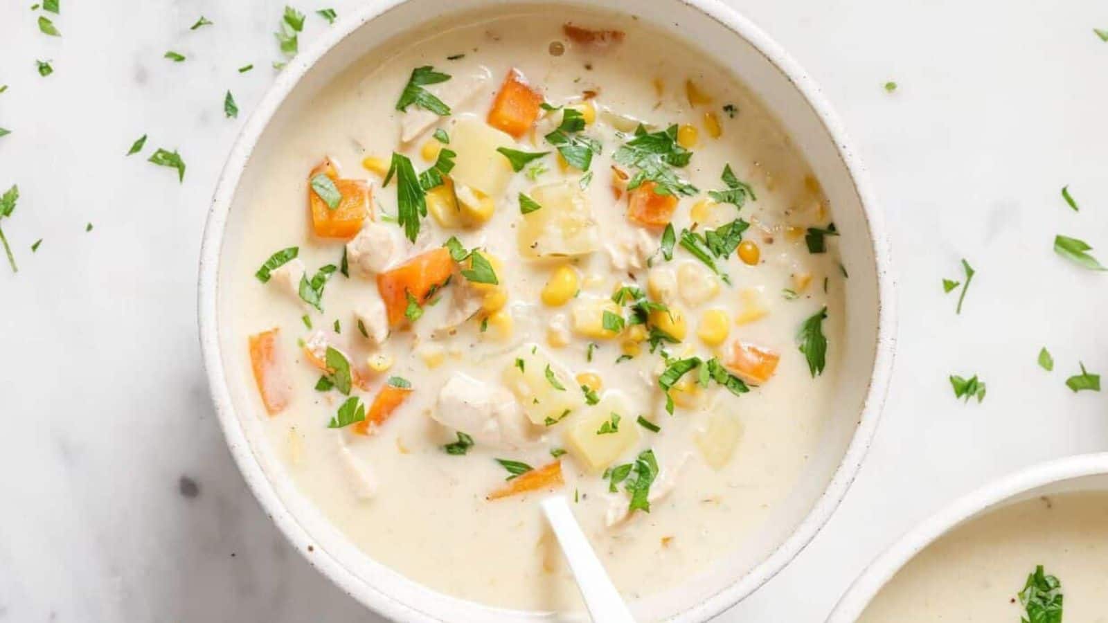 A bowl of creamy chicken soup containing diced carrots, corn, potatoes, and herbs. The soup is garnished with chopped parsley and has a white spoon in it. The bowl is placed on a light-colored surface.