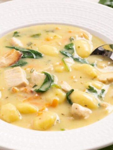 A bowl of creamy soup containing gnocchi, chicken pieces, and spinach, garnished with herbs. A spoon is placed inside the bowl, resting on the rim. The bowl is on a white plate on a wooden surface.