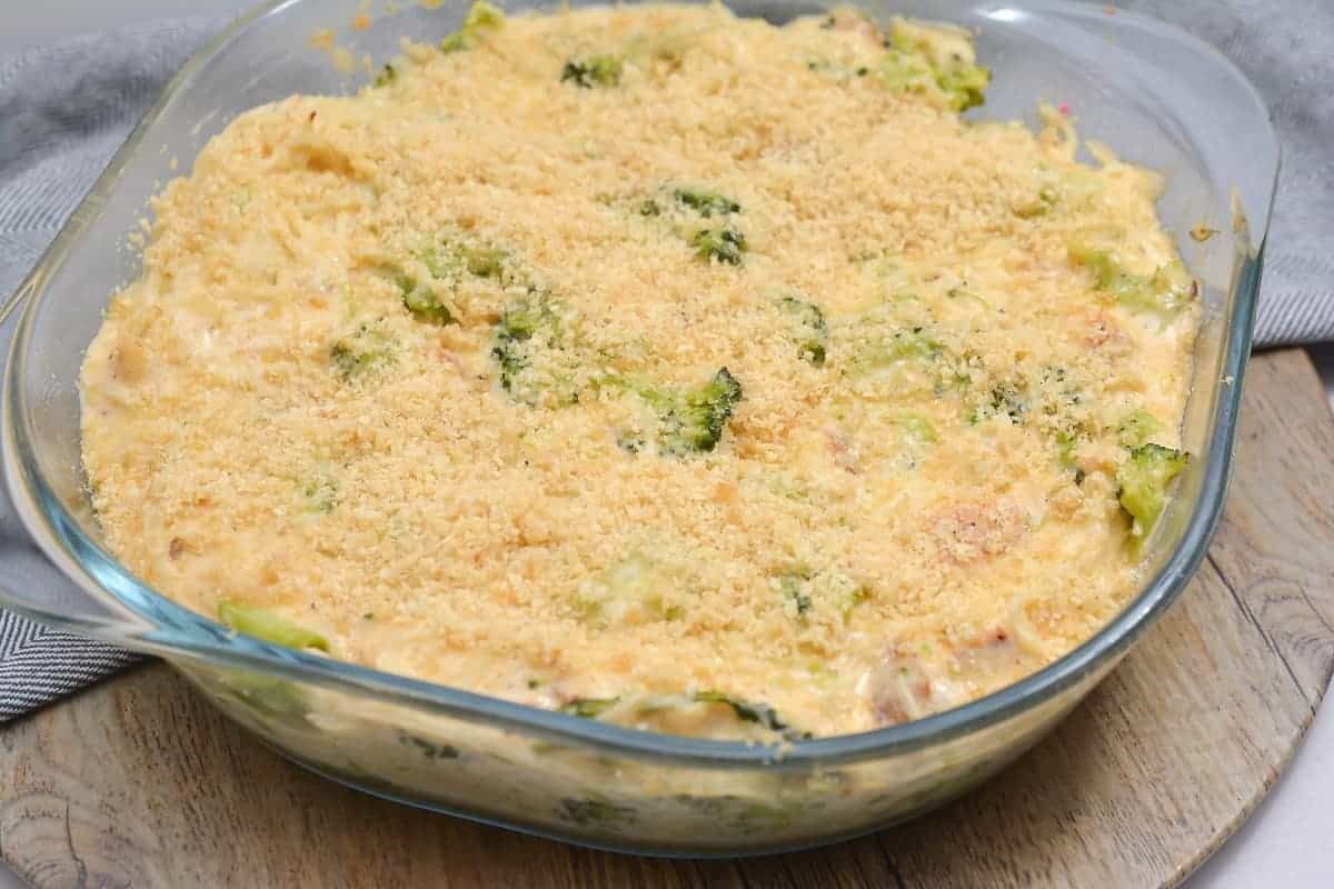 A glass baking dish filled with a baked casserole, an ideal all-in-one meal for effortless weekends, topped with breadcrumbs. The casserole boasts visible pieces of broccoli mixed into a creamy sauce. It rests on a wooden surface with a gray cloth nearby, promising comfort and simplicity.