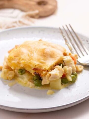 A plate featuring a serving of chicken pot pie with a golden-brown crust and visible pieces of vegetables, ideal for hearty meals. A fork sits on the side, and a glass of orange juice is partially visible in the background, making it perfect for big gatherings.