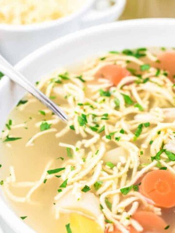 A bowl of chicken noodle soup contains shredded chicken, noodles, and sliced carrots, garnished with chopped parsley. A spoon rests in the bowl. Fresh parsley and a bowl of grated cheese are in the background.