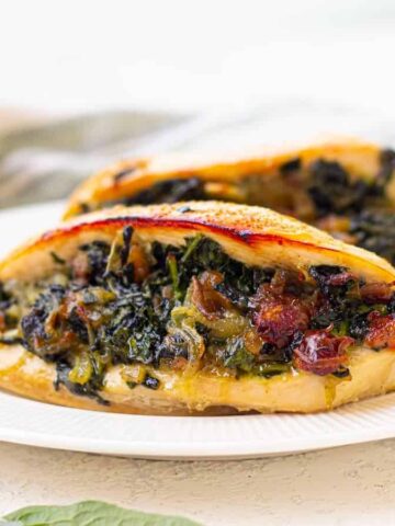 Two pieces of stuffed chicken breast are on a white plate. The chicken is filled with a mixture of spinach, bacon, and cheese. A striped napkin and a few spinach leaves are in the background on a light-colored surface.