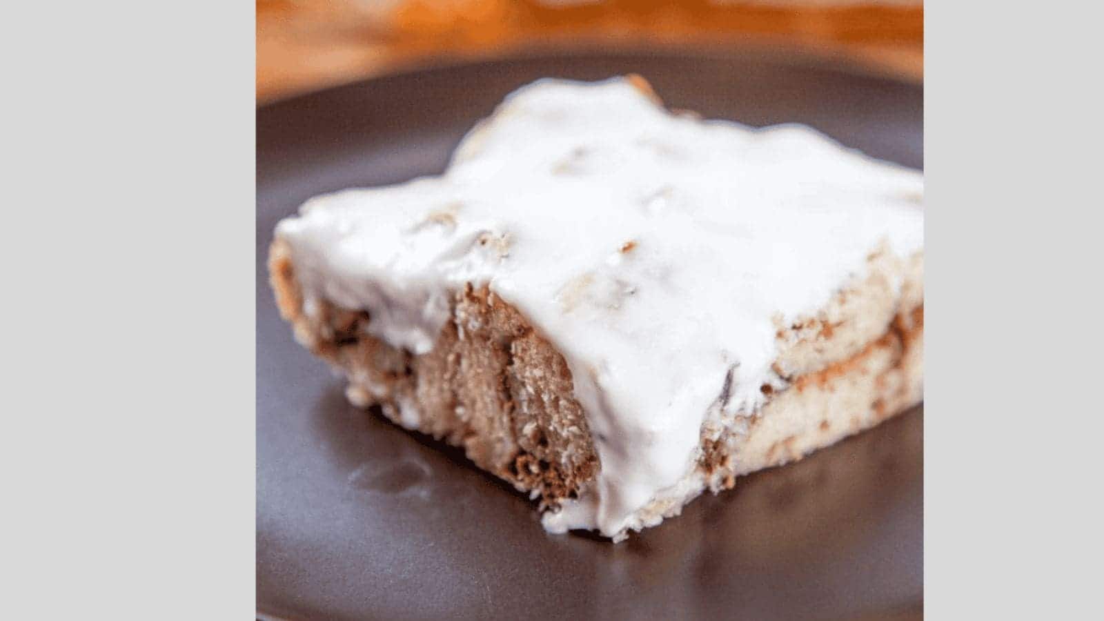 A piece of cake with white frosting on a dark plate. The cake appears to have a light, fluffy texture and is topped evenly with the frosting. The background is softly blurred.