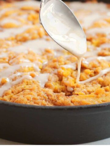 A spoon drizzles glaze over a freshly baked sweet potato dessert in a black skillet, showcasing its golden-brown, crumbly texture.