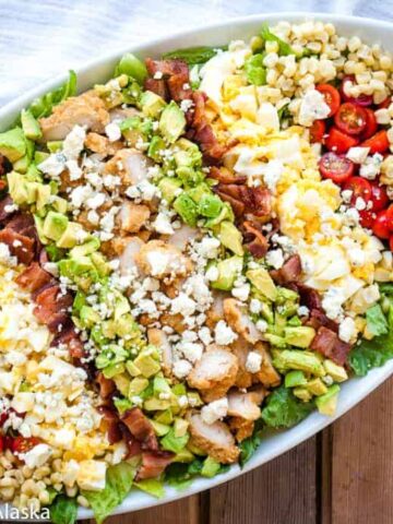 A large oval platter is filled with a Cobb salad, perfect for sharing with loved ones. It features sections of chopped grilled chicken, diced avocado, cherry tomatoes, corn kernels, crumbled cheese, bacon pieces, and hard-boiled eggs, all arranged over a bed of mixed greens.