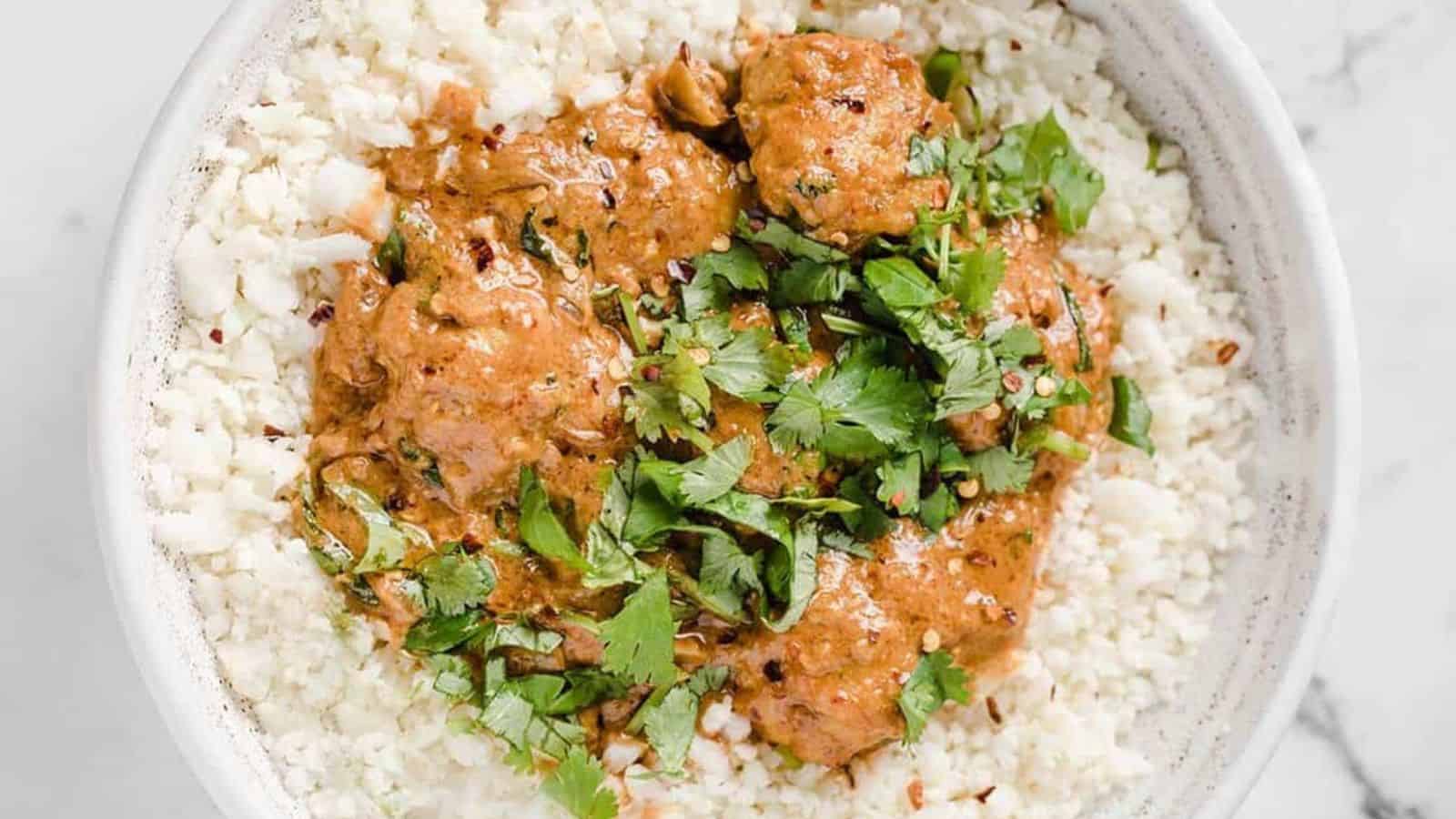 A bowl of cauliflower rice topped with a creamy tomato-based curry and garnished with fresh cilantro. The dish is served in a white bowl on a white surface.