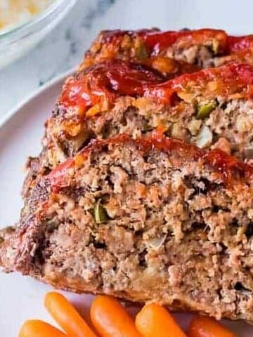 Slices of meatloaf topped with a glaze are served on a white plate alongside mashed potatoes and steamed baby carrots. A bowl with a portion of the meal is partially visible in the background.