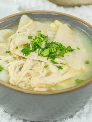 A bowl of chicken soup, an epitome of effortless weekends, is served with shredded chicken, potatoes, and chopped fresh parsley. A silver spoon rests in the bowl on a white textured surface, with a few parsley leaves adding to the cozy scene.