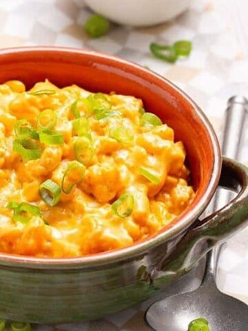 A ceramic bowl filled with creamy mac and cheese, garnished with chopped green onions. A spoon rests beside the bowl on a checkered napkin. The setting includes a portion of a pot and a small white dish in the background.
