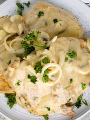 A plate of creamy, no-fuss smothered pork chops topped with sliced onions and mushrooms, garnished with parsley. Perfect for lazy weekends, a striped cloth is partially visible on the right side of the gray stone surface.