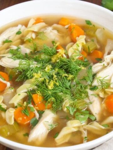 A bowl of chicken noodle soup featuring chunks of chicken, sliced carrots, celery, and pasta noodles in broth. Fresh dill and parsley are sprinkled on top. The bowl is placed on a wooden surface with herbs and a spoon nearby.