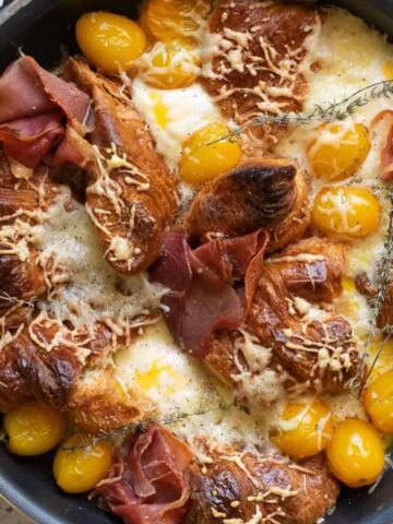 A skillet filled with baked croissants, melted cheese, yellow cherry tomatoes, and slices of cured meat, garnished with fresh thyme. A grey cloth is draped under the skillet on a speckled countertop.