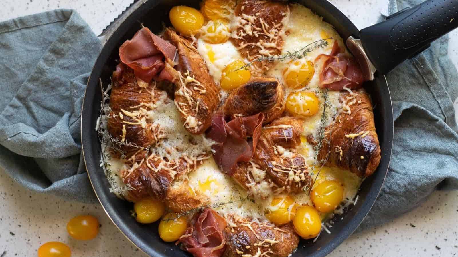 A skillet filled with baked croissants, melted cheese, yellow cherry tomatoes, and slices of cured meat, garnished with fresh thyme. A grey cloth is draped under the skillet on a speckled countertop.