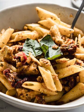 A white bowl filled with penne pasta mixed with chunks of chicken, sun-dried tomatoes, and garnished with fresh basil leaves. A fork is placed in the bowl, and a small dish is visible in the background.