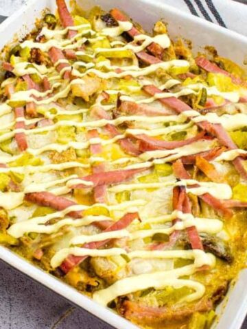 A square baking dish filled with a baked casserole topped with strips of ham and drizzles of creamy sauce. The casserole appears to contain sliced vegetables and melted cheese. A striped cloth is partially visible beside the dish.