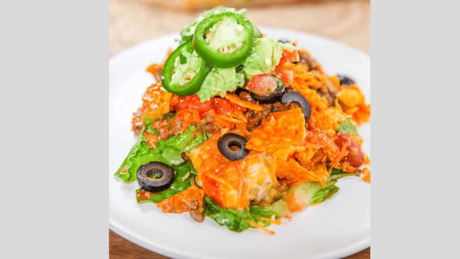 A plate of loaded nachos topped with jalape&ntilde;o slices, avocado, black olives, tomatoes, melted cheese, and lettuce. The nachos are layered with crispy chips and garnished with a spicy kick.