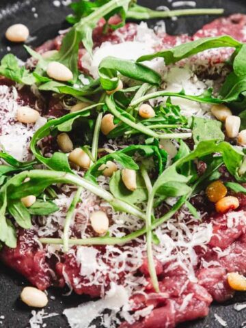 A black plate features slices of raw beef topped with arugula, grated cheese, and pine nuts. The dish rests on a light-colored tabletop.