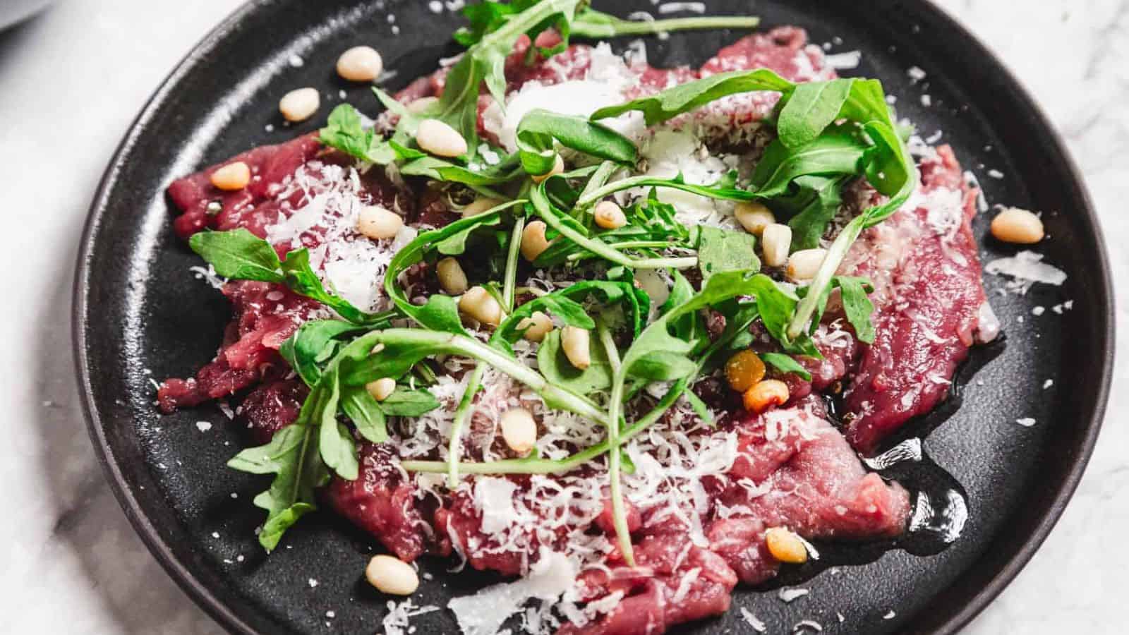 A black plate features beef carpaccio topped with arugula, grated Parmesan cheese, and pine nuts. The thinly sliced meat is arranged with the garnishes spread evenly across its surface.