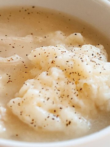 A bowl of soup features pieces of shredded chicken and dumplings, with the surface speckled with black pepper. The soup is light in color, suggesting a broth-based preparation. The bowl is white and placed on a white plate.