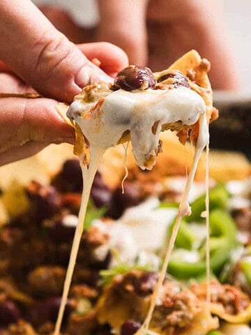 A hand is holding a tortilla chip covered with melted cheese, ground meat, and beans. Below, other chips are topped with similar ingredients, along with sliced green onions, creating a tray of nachos.