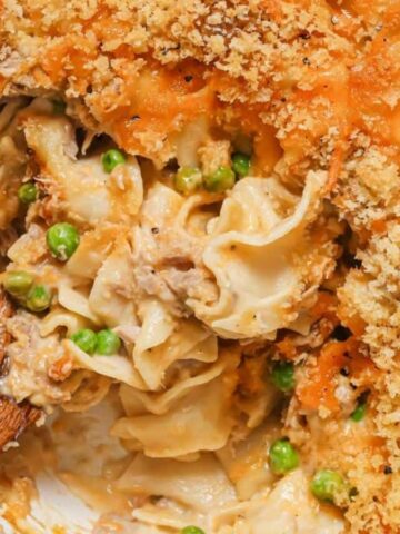 A casserole dish filled with a creamy noodle and vegetable mixture, topped with a golden breadcrumb crust. A wooden spoon is placed in the dish, showing a portion of the creamy filling containing peas and chunks of meat.