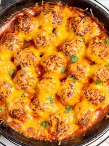 A skillet filled with meatballs covered in melted cheese and sauce sits enticingly on a white surface. For those cozy evenings, it’s garnished with chopped herbs, with a bowl of herbs and fresh cilantro nearby, making it the perfect savory oven meal.