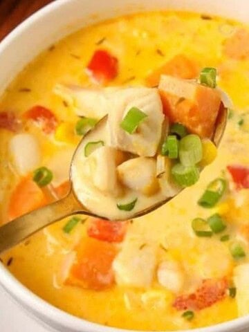 A bowl of creamy corn chowder with chunks of vegetables and garnished with chopped green onions. A spoon is held above the bowl, showing a close-up of the chowder. Sliced bread and a small bowl of additional green onions are in the background.