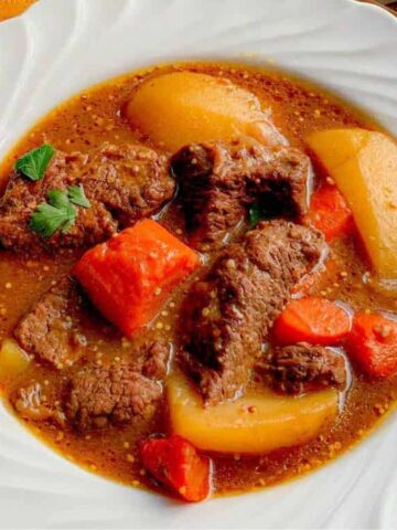 A white bowl cradles a hearty beef stew with tender chunks of beef, potatoes, and carrots in a rich brown broth. Perfect for winter meals, it's garnished with a sprig of parsley. An orange napkin and a spoon are placed beside it, making it an inviting dish everyone would recommend.