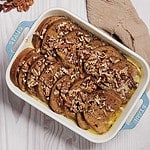 A rectangular baking dish filled with a decadent pecan praline French toast casserole, topped with chopped pecans. The dish sits on a light wooden surface with a beige cloth to the side. The bread is soaked in a rich, visible egg mixture.