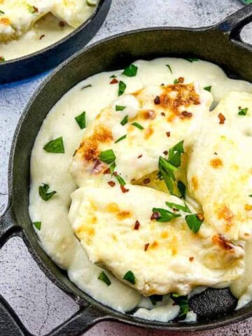 Two small cast iron skillets contain creamy mashed potatoes topped with melted cheese and herbs, channeling your inner grandma's comfort foods. They rest on a textured gray surface, with a patterned cloth napkin partially visible to the right.