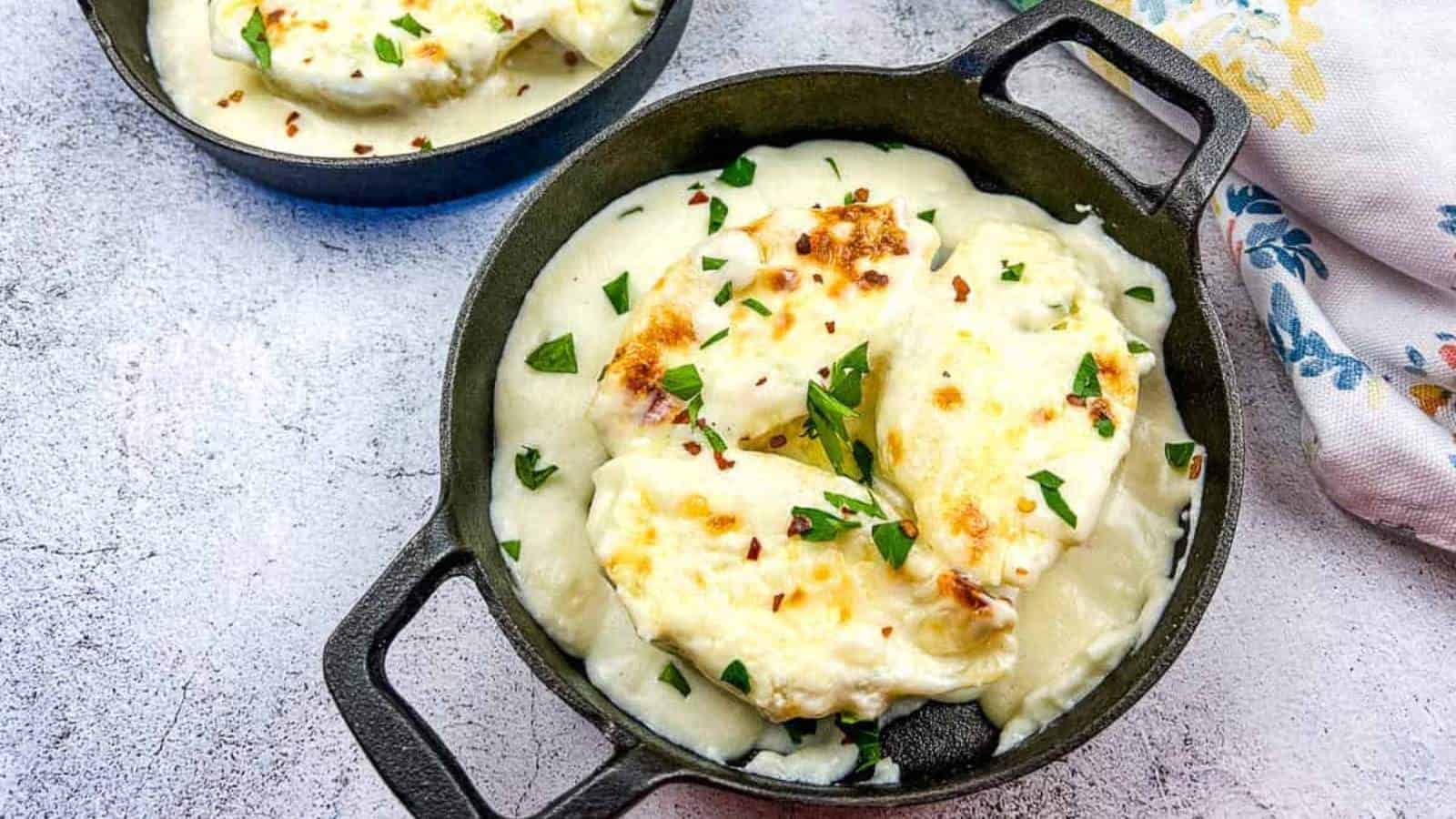 Two small cast iron skillets contain creamy mashed potatoes topped with melted cheese and herbs, channeling your inner grandma's comfort foods. They rest on a textured gray surface, with a patterned cloth napkin partially visible to the right.