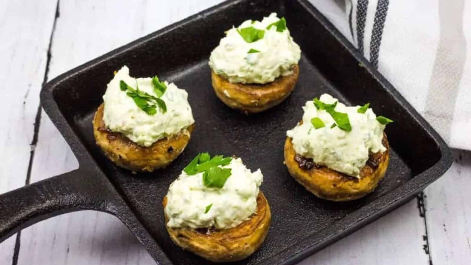A black square skillet contains four stuffed mushrooms topped with a creamy white mixture and garnished with chopped parsley. Perfect for sharing during family recipes or gatherings with friends, the skillet rests on a white wooden surface, while a striped cloth peeks in from the top right corner.