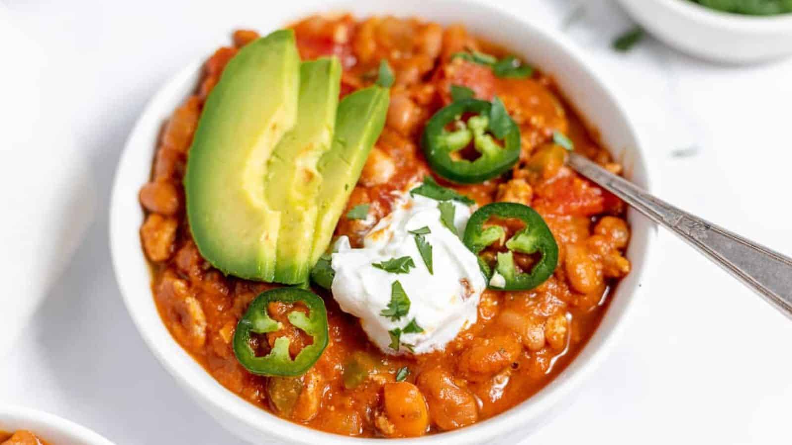A bowl of chili topped with sliced avocado, jalape&ntilde;os, and a dollop of sour cream. There is a spoon in the bowl. The chili is garnished with chopped cilantro.