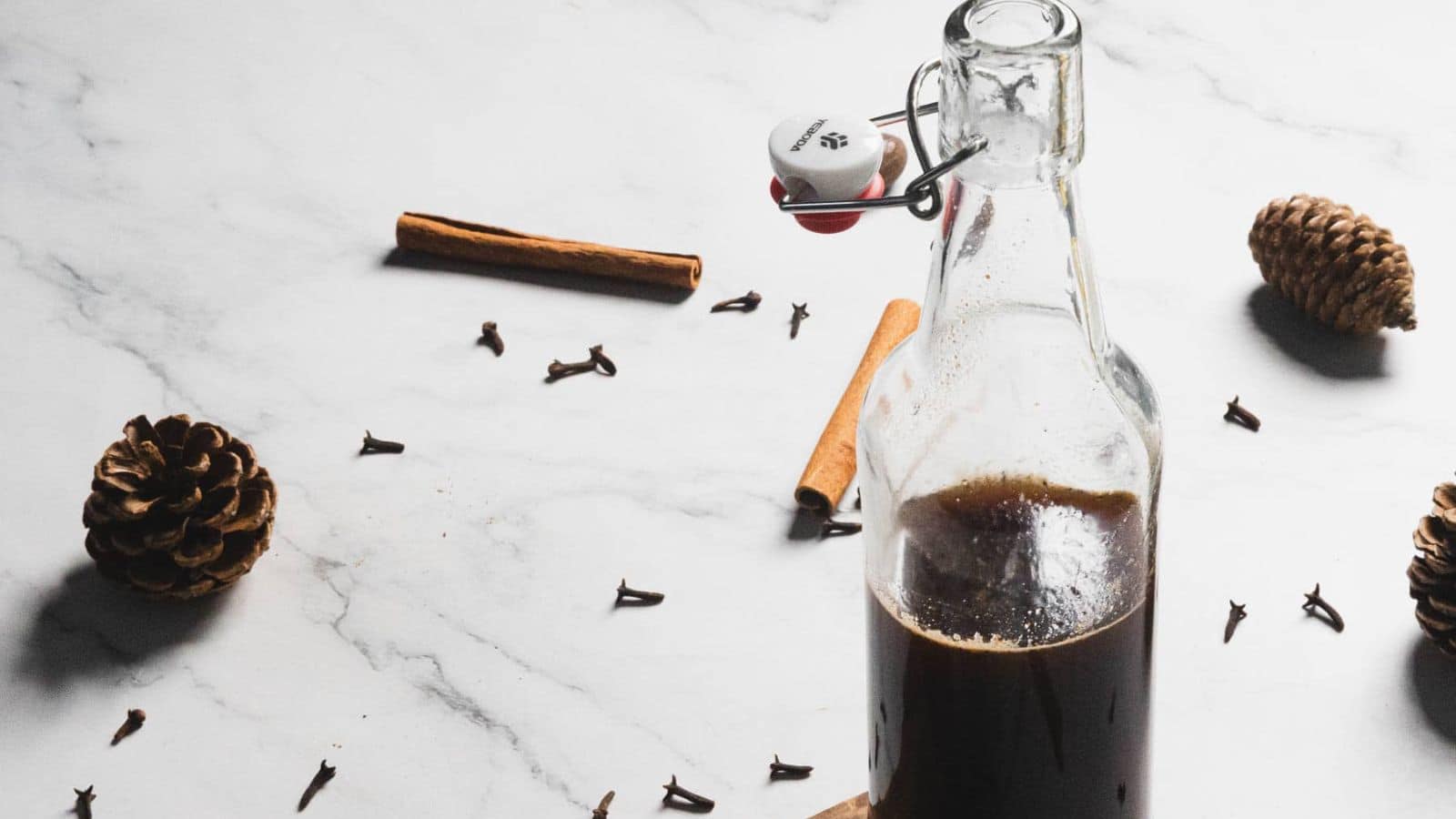 A glass bottle containing a dark brown liquid sits on a marble surface. Around it are scattered cinnamon sticks, cloves, and pinecones, creating an autumnal theme. A metal clasp with a white stopper seals the bottle.