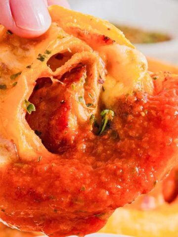 A close-up of a hand holding a pizza roll. The roll is covered in melted cheese, tomato sauce, and herbs. Other pizza rolls are visible in the background on a white surface.