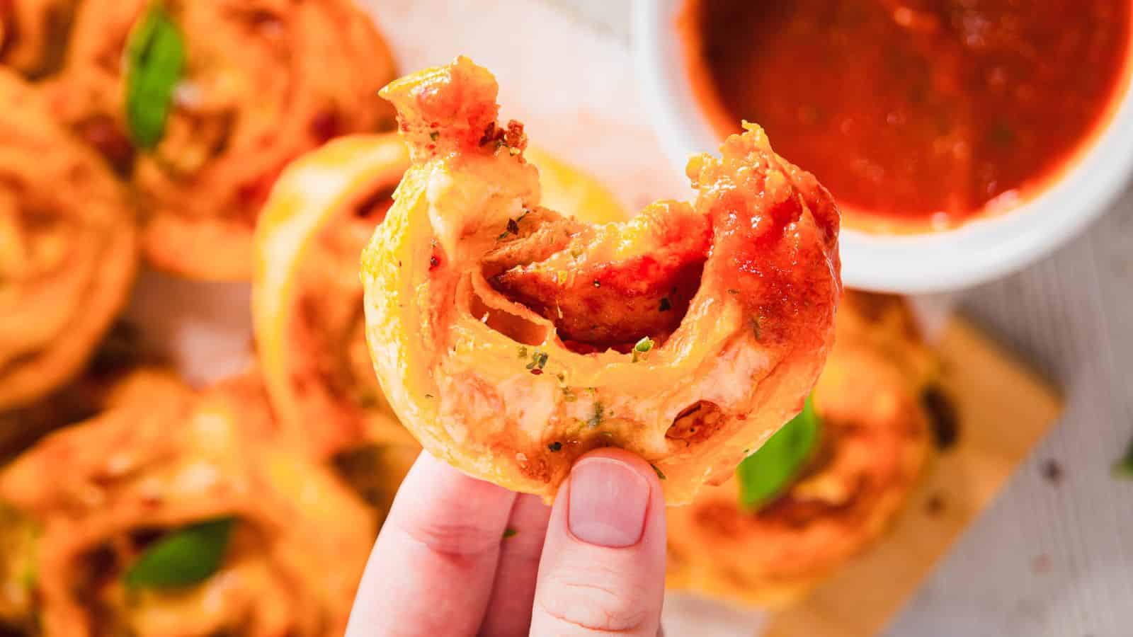 A hand holding a piece of pastry roll topped with melted cheese, herbs, and tomato sauce. A bowl of red dipping sauce is in the background, placed on a wooden surface with more pastry rolls.
