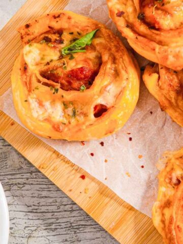 A wooden board holds several spiral-shaped pastry rolls filled with cheese and tomato, garnished with basil. A small bowl containing red sauce is placed to the side. The background features a textured grey surface.