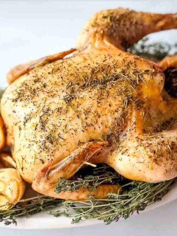 A roasted chicken seasoned with herbs sits on a white platter, surrounded by sliced lemons, roasted garlic, and fresh sprigs. This oven meal on a white marble surface offers a taste of relaxing cooking with easy recipes for any occasion.